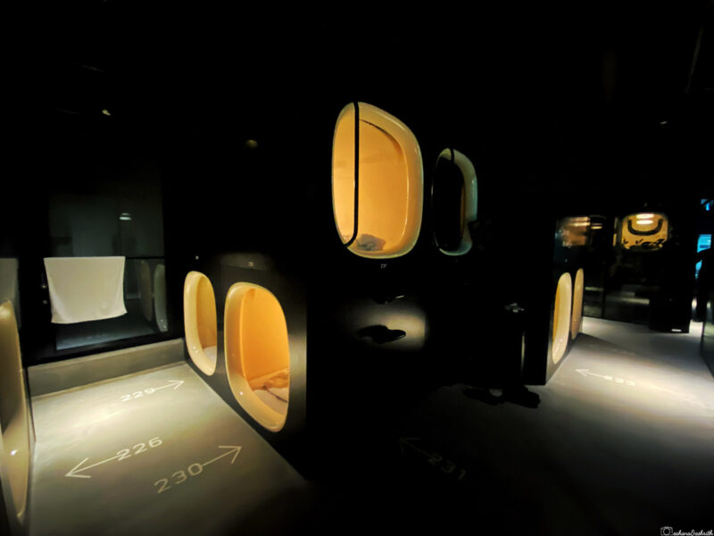 A dark, futuristic capsule hotel room with illuminated sleeping pods in a row.