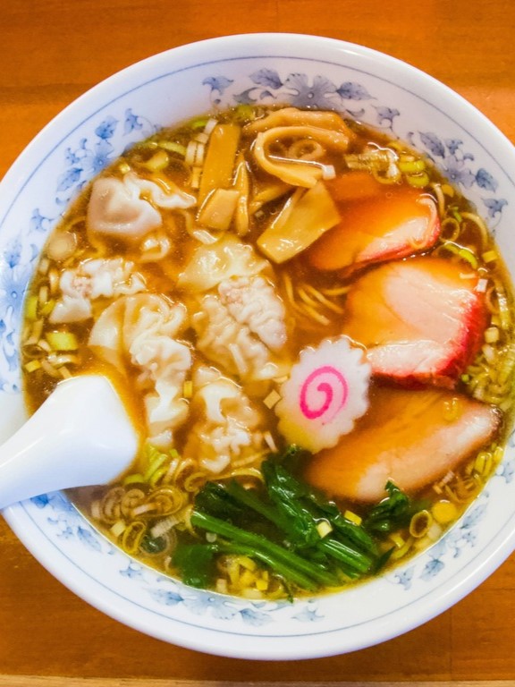 Bowl of ramen topped with sliced pork, bamboo shoots, seaweed, and fish cake, with a spoon on the side.