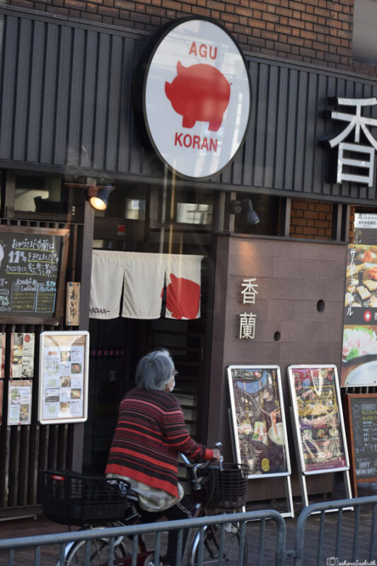  Exterior of a restaurant named Agu Koran with a large pig logo, an elderly woman cycling past