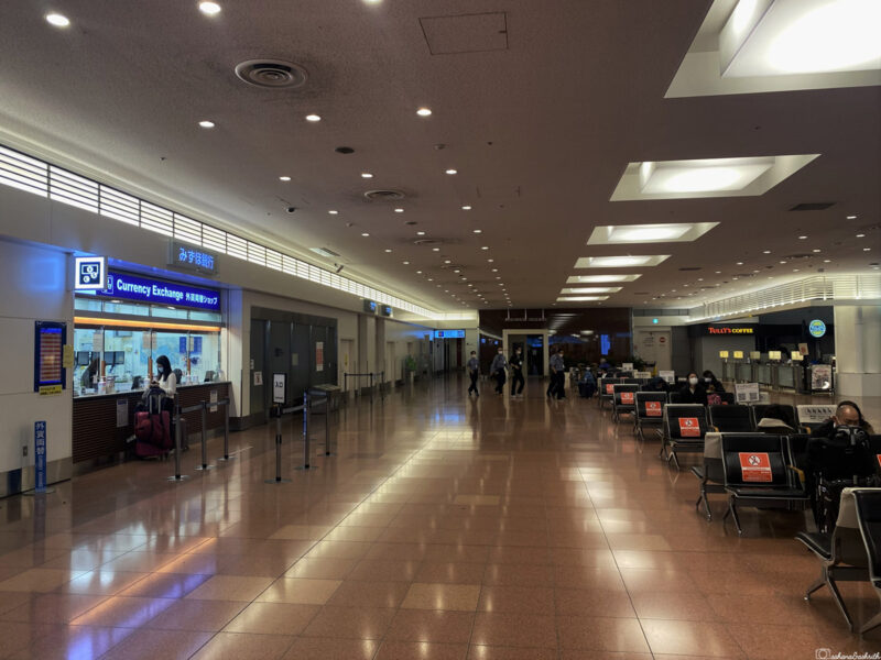 Empty airport corridor with a currency exchange booth and seating area, illuminated by overhead lights.