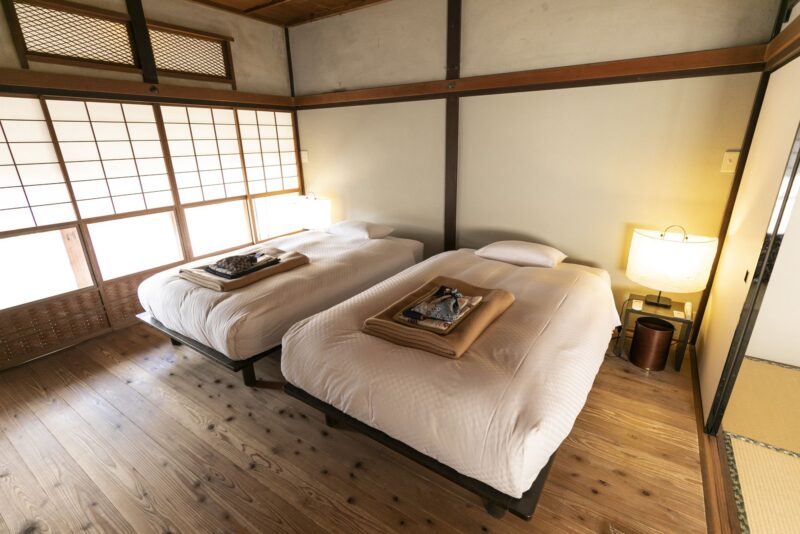 Simple Japanese-style bedroom with two single beds, soft lighting, and shoji screens on wooden flooring.