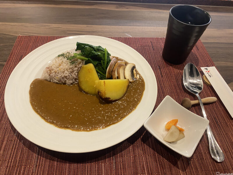 Plate of Japanese curry rice with spinach, mushrooms, and potatoes, accompanied by pickles and a drink.