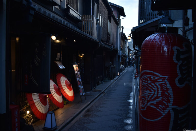 Narrow alleyway in Kyoto lined with traditional Japanese shops and large red lanterns glowing in the evening.