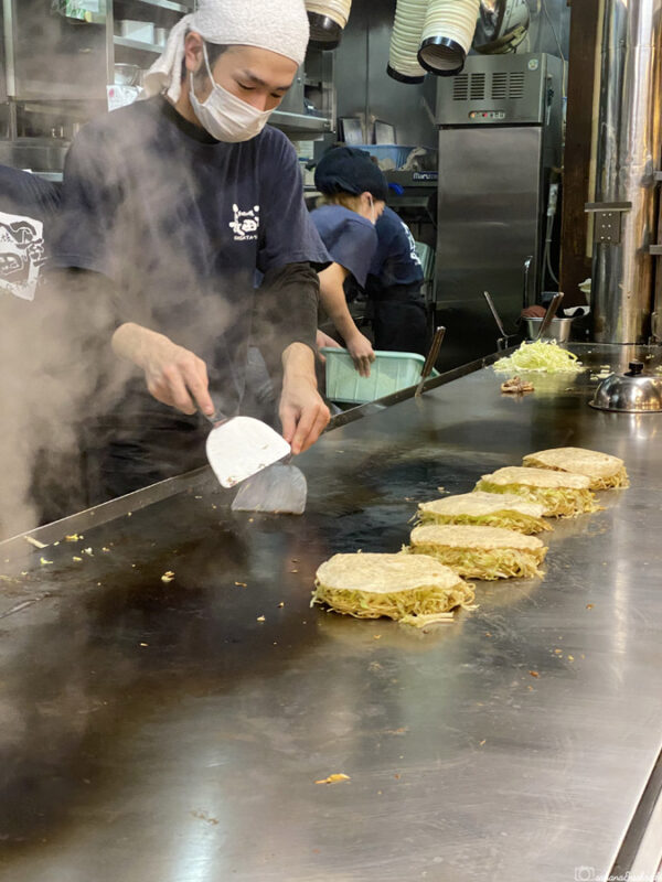 Chef grilling multiple okonomiyaki pancakes on a hotplate in a smoky kitchen.