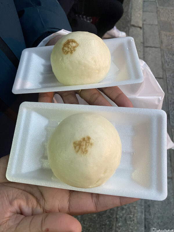 Two steamed buns on white foam trays, each held by a person's hand, visible on a grey street