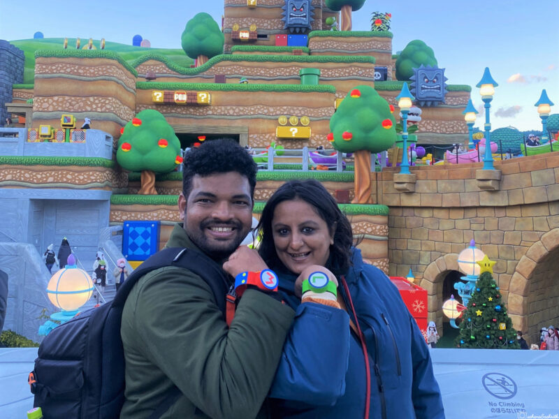 A couple smiling at Super Nintendo World, wearing colorful wristbands in front of a bright, whimsical landscape