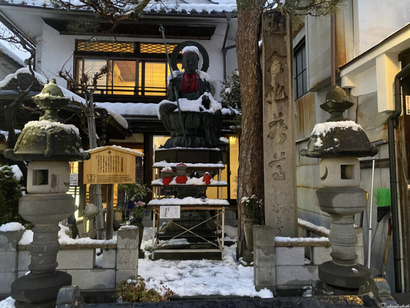 Snow-covered statue of a seated Buddhist figure, flanked by two stone lanterns, outside a traditional Japanese building