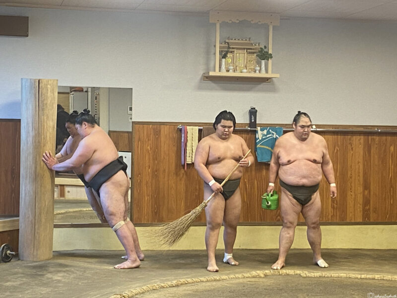 One sumo wrestler practices teppo exercises while two others clean the ring with a broom and watering can.