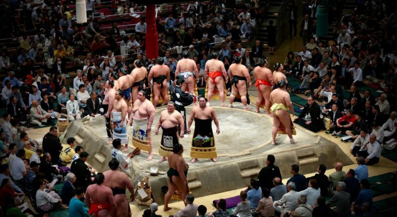 Sumo wrestlers in ceremonial attire gather in the ring, facing the audience during a sumo tournament