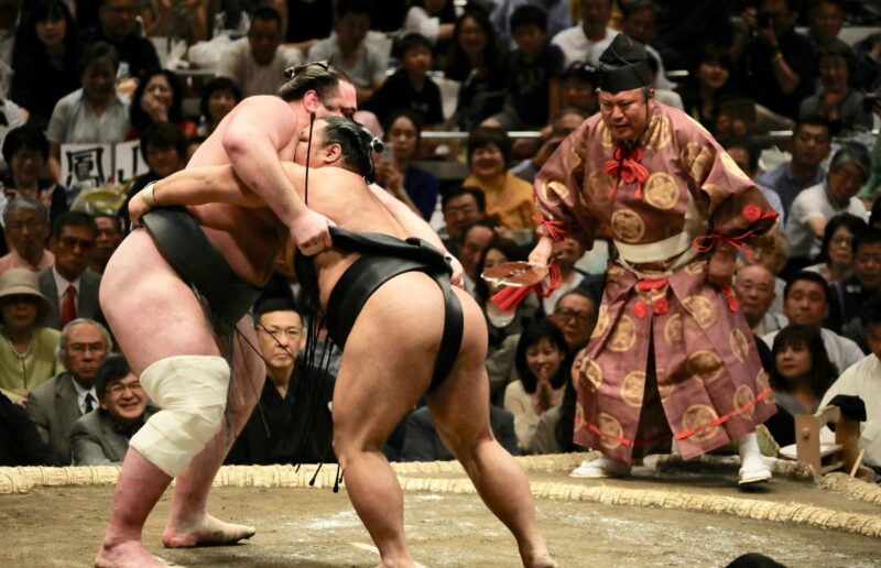 Two sumo wrestlers grapple in a ring while a traditional referee watches closely, with a large audience observing the intense match.