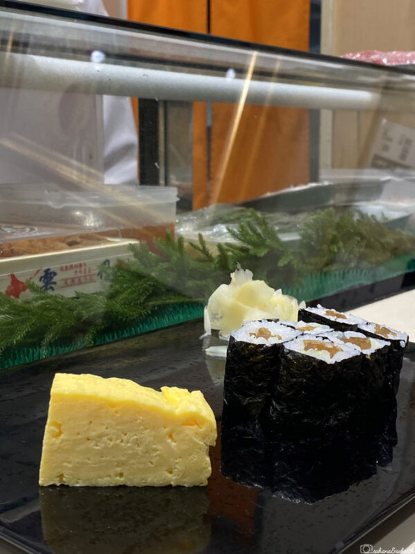 Sushi counter with tamagoyaki, maki rolls, and a slice of pickled ginger on a black plate