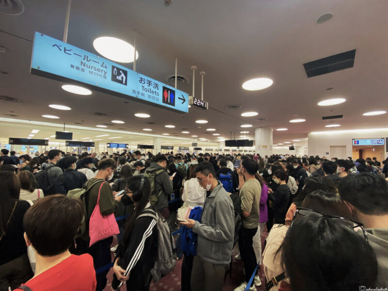 Crowded haneda airport terminal with travelers waiting under a sign pointing towards the nursery and toilets.