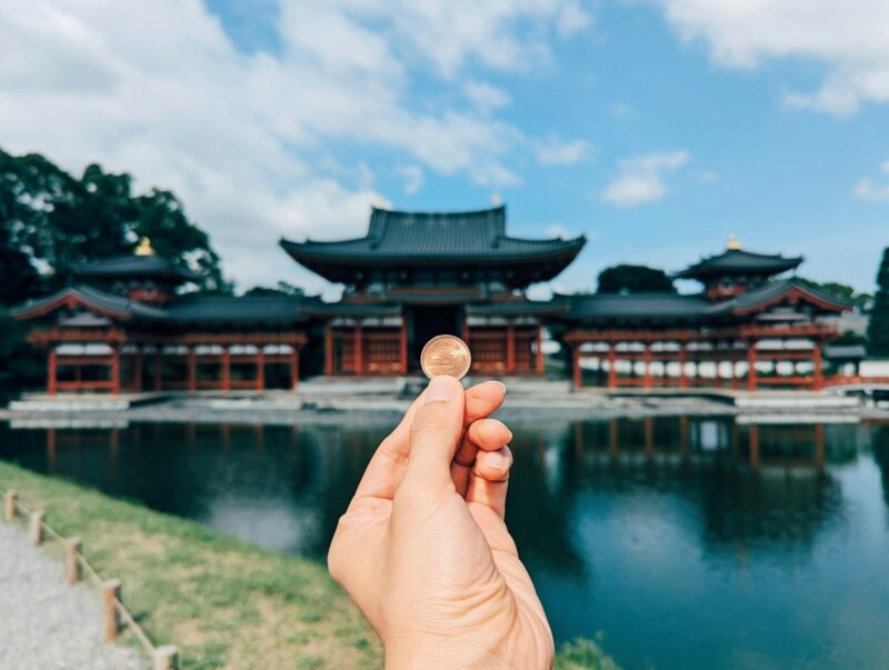 A hand holding a ten coin in front of the Byodo-in Temple in Uji, Japan, reflecting in the calm water.