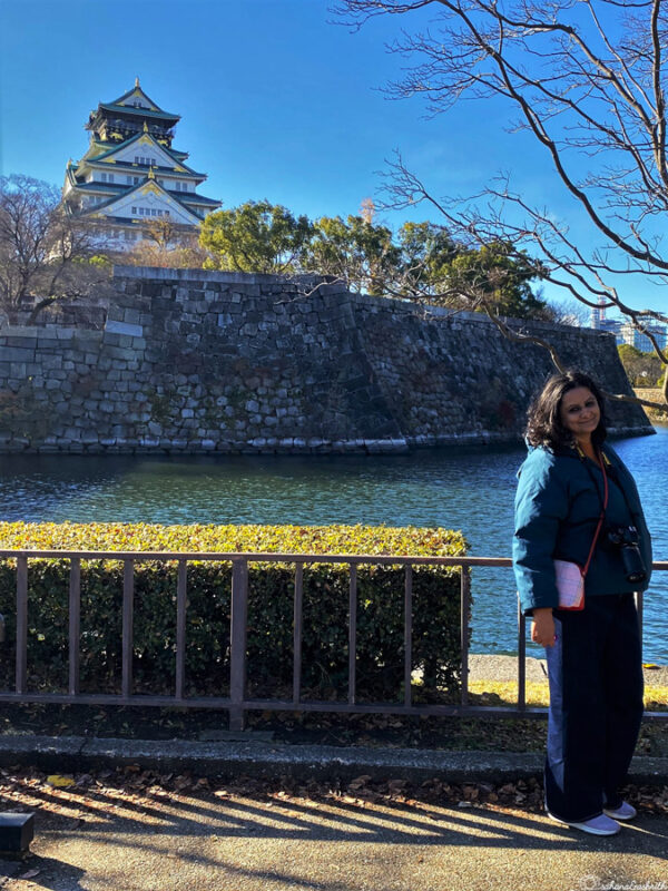 Woman smiling by the moat with Osaka Castle in the background on a sunny day, surrounded by trees and clear blue skies.