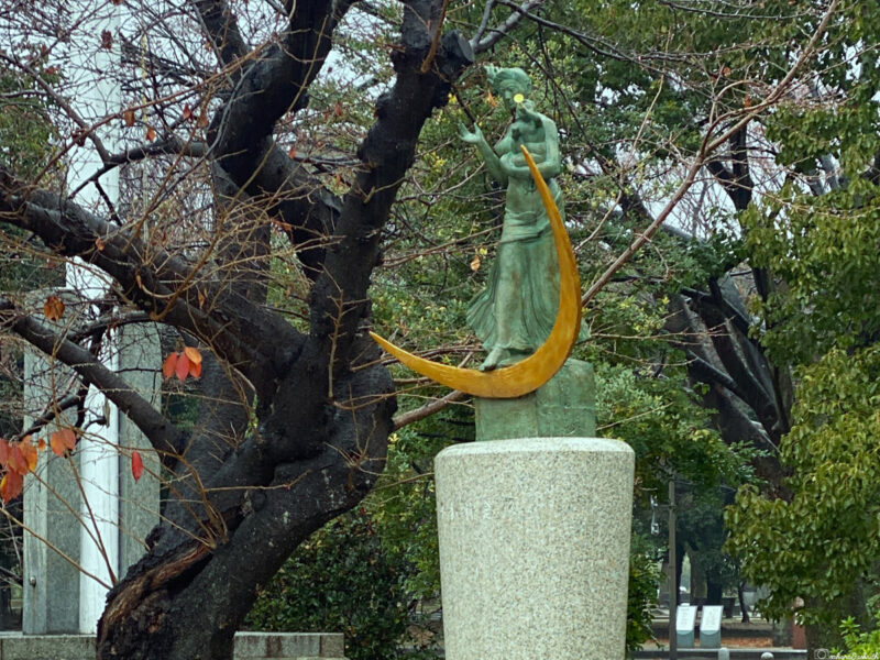 A bronze statue of a woman holding a golden crescent moon, surrounded by autumn trees, located in Hiroshima Peace Memorial Park, symbolising resilience and memory.