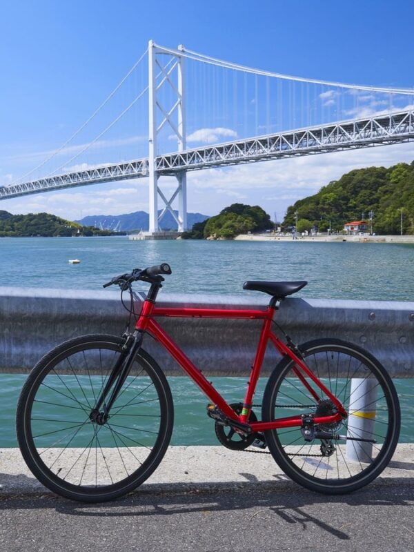 Red bicycle parked near Seto Inland Sea with the Great Seto Bridge towering in the background under a clear blue sky, calm water beneath bridge