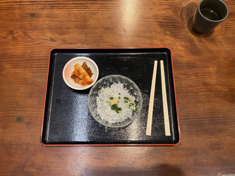 tray with a bowl of kamakura famous whitebait (shirasu) topped with green onions, a side of pickled vegetables, and chopsticks, served in a traditional Kamakura restaurant