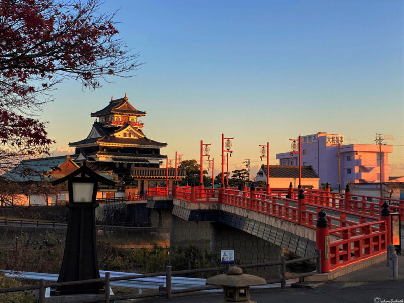 sunset view of Kiyosu Castle with red bridge lanterns and a foreground framed by a lamp post and autumn leaves. (