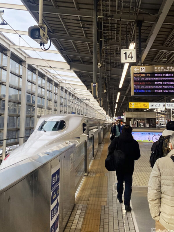 hinkansen platform in Hiroshima station, sleek white bullet train preparing to depart as passengers stand nearby, destination signs visible