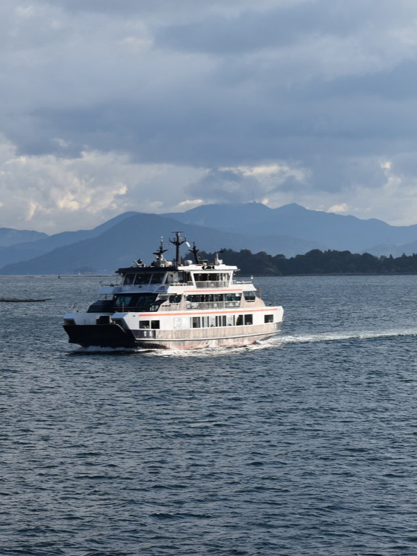 A ferry cruising through calm waters near Hiroshima, with mountainous islands visible in the background under a partly cloudy sky.