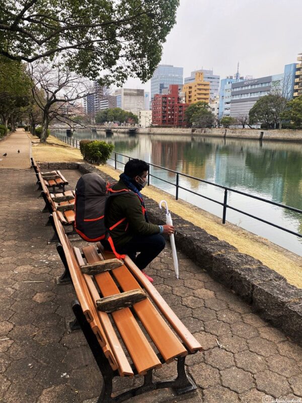 Traveller sitting on a riverside bench in Hiroshima with urban skyline in the background, taking a moment to relax by the water