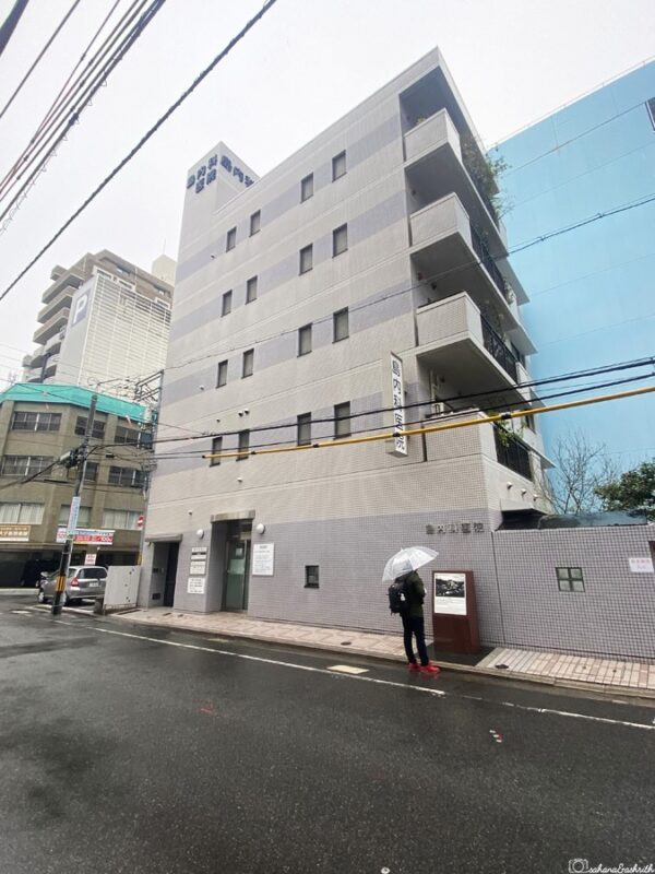 Modern grey shima clinic building in Hiroshima with visitor holding umbrella outside, illustrating simple budget accommodation option in the city