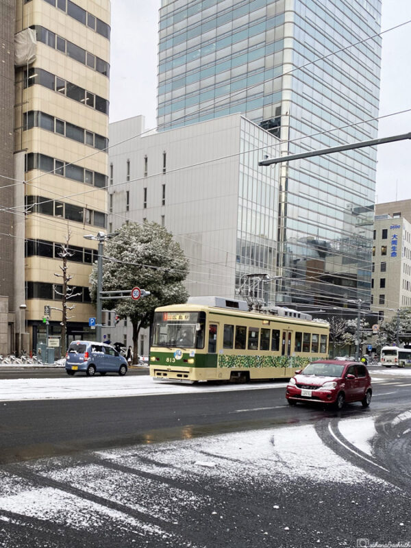 City tram in Hiroshima covered in green leaf design, moving through snowy streets among modern buildings with light snow dusting road and trees