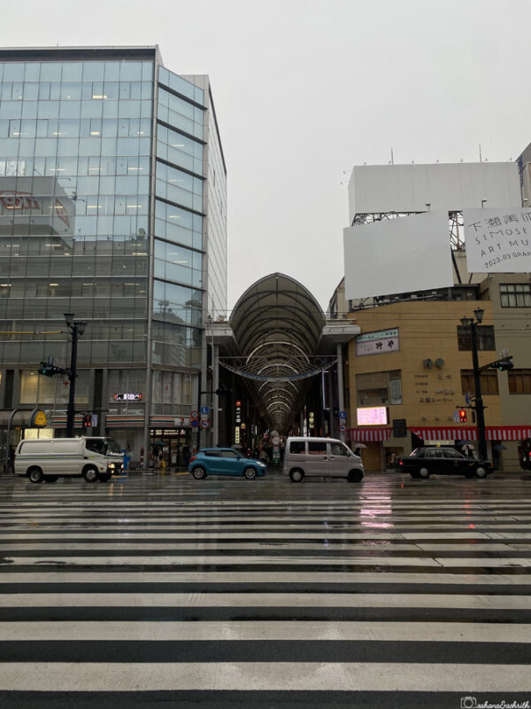 Covered shopping arcade in Hiroshima with cars crossing at the zebra crossing, surrounded by modern glass buildings on a rainy day