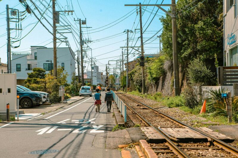A quiet street in Kamakura runs alongside railway tracks, with two kids strolling under the sun, capturing the town's peaceful vibe
