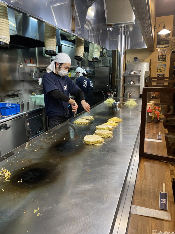 Nagataya restaurant chef preparing okonomiyaki on a teppan grill, creating popular Japanese savoury pancakes in a lively local setting