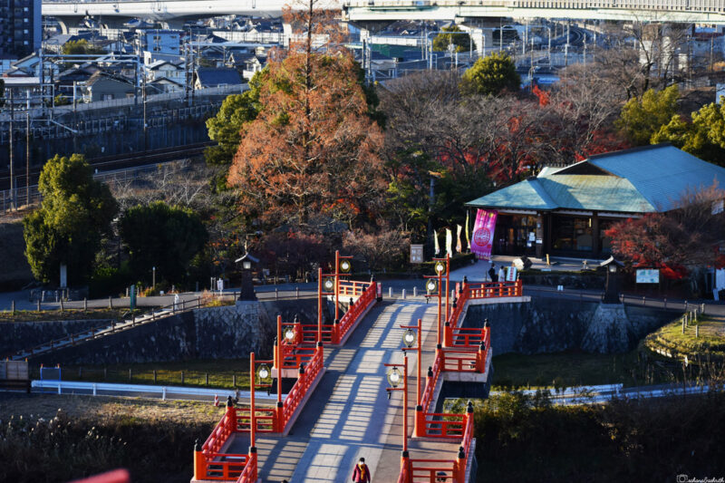 Aerial view of a vibrant red otebashi bridge with a castle in the distance, surrounded by trees and city infrastructure in the background
