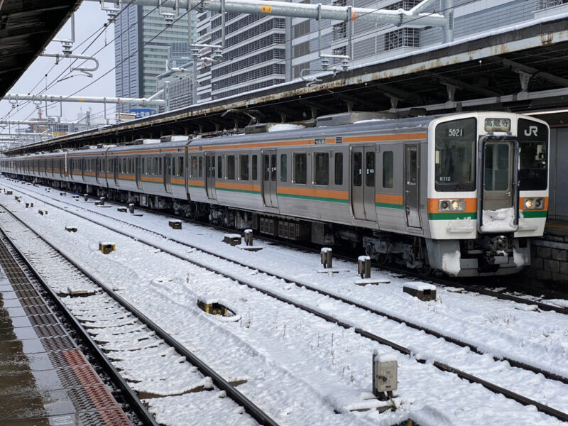 A snowy Nagoya JR train station featuring a long orange and green train parked on tracks, surrounded by modern urban buildings