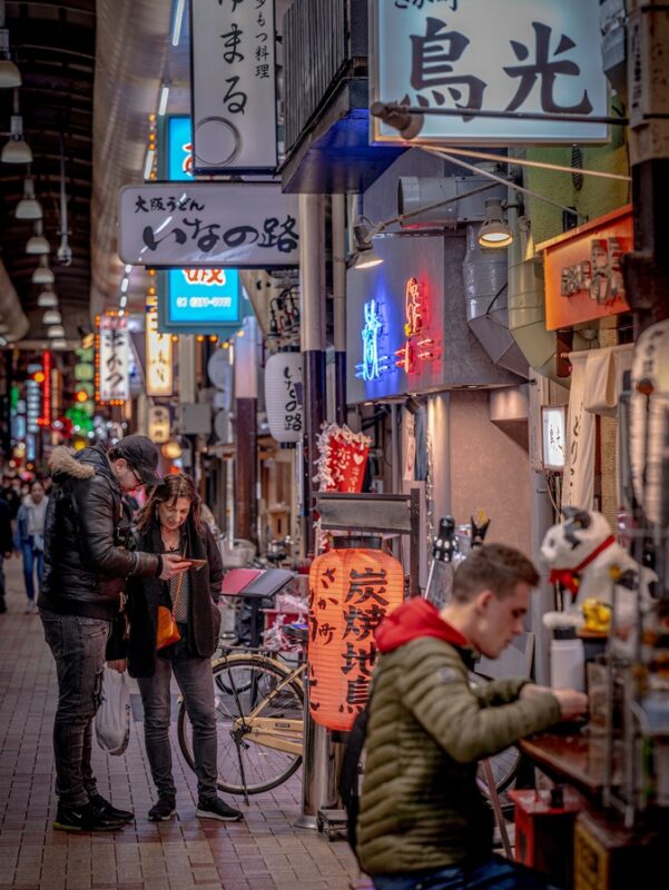 Busy food alley in Osaka at night with illuminated signs and people exploring local eateries, capturing the city’s vibrant nightlife.