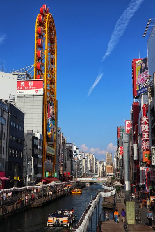 Bright, bustling Dotonbori canal in Osaka with boats, colourful buildings, and the iconic Ferris wheel under a clear blue sky