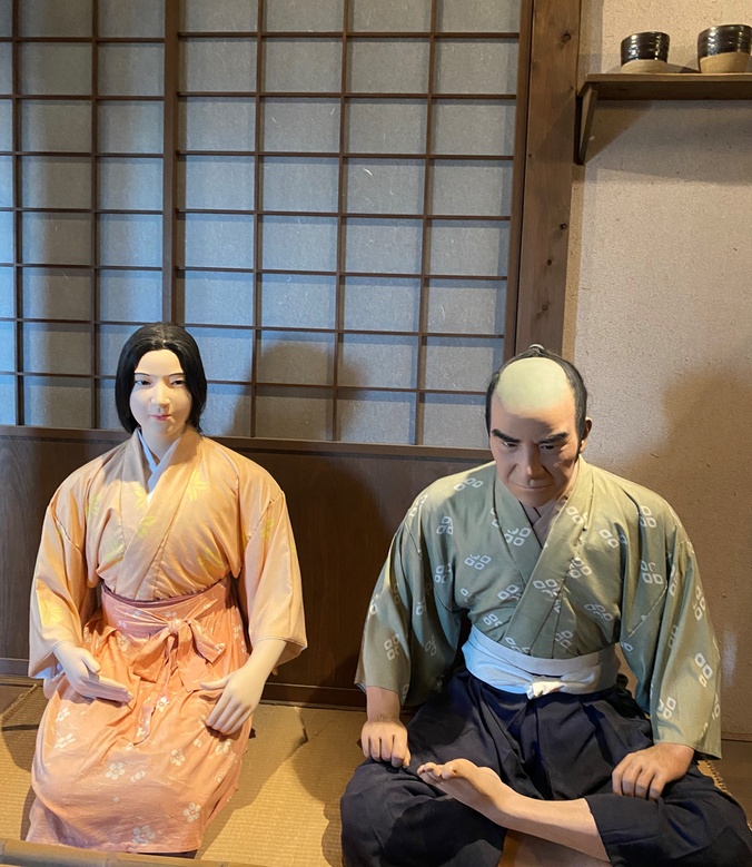 Mannequins of samurai couple dressed in traditional Japanese attire, seated on tatami mats inside a room with shoji screens, representing historical Japanese life.