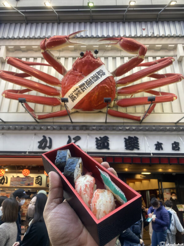 Hand holding sushi box in front of the famous giant crab sign at Kani Doraku restaurant in Dotonbori, Osaka.