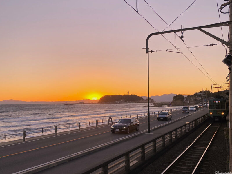 A stunning sunset along the coast in Kamakura, with cars and a train travelling parallel to the glowing horizon over the sea at kamakura kokomae station