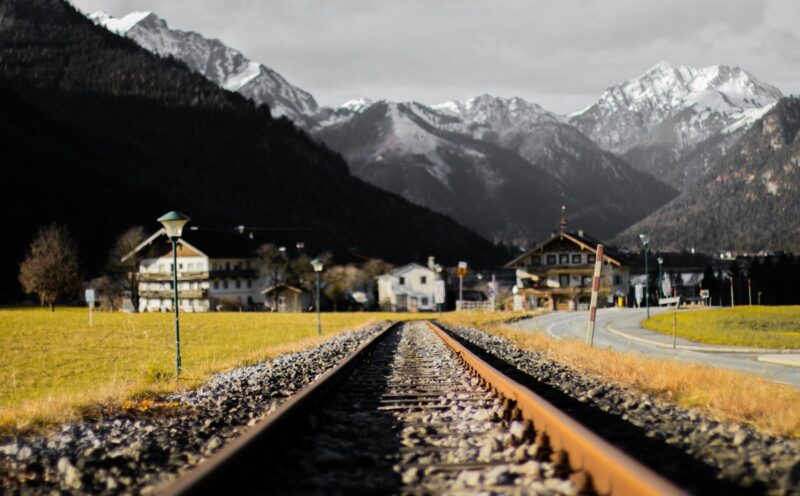 Rusty railway tracks lead toward snow-capped mountains with village houses in the Austrian Alps.