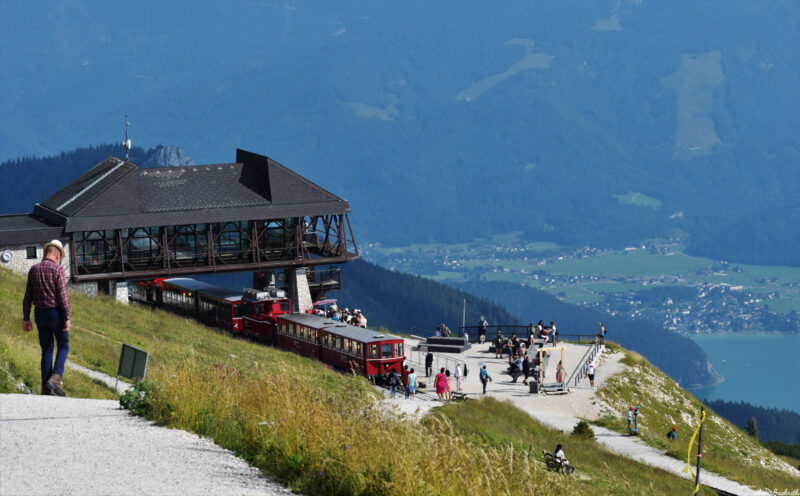 Beautiful Schafberg peak with a train station, turquoise lake at the bottom, and hikers climbing further up.
