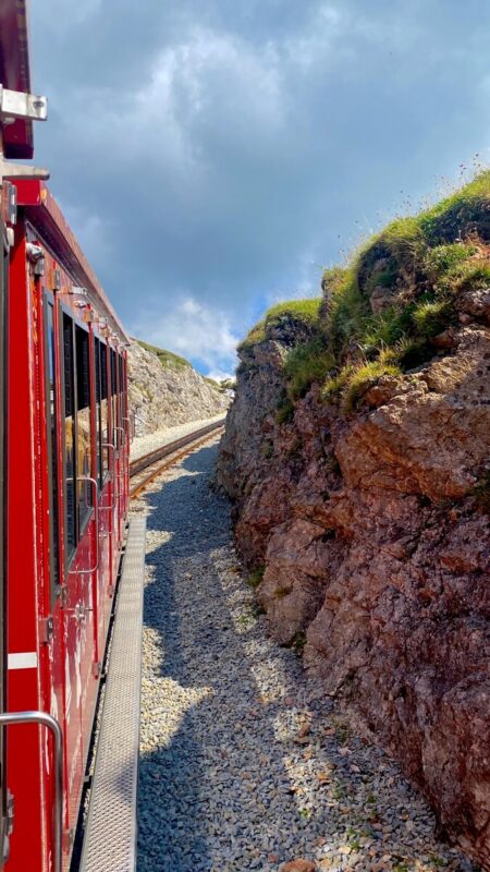 A red train travels through rocky terrain on the steep tracks of the Schafberg Railway.