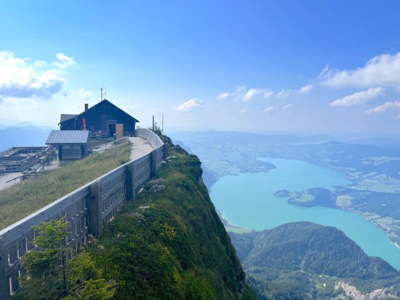 A wooden cabin sits on a cliff with views of turquoise Lake Wolfgangsee and green hills at Schafbergspitz