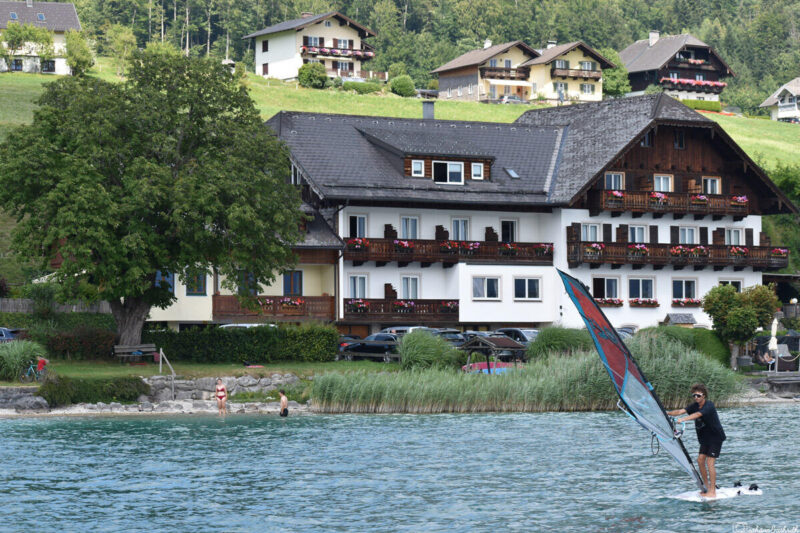 A kayaker on Wolfgangsee lake on a sunny afternoon, with a beautiful house adorned with balcony flowers and meadows along the bank.