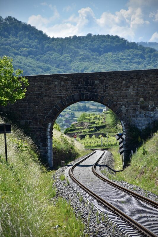 A stone railway arch frames tracks with green hills and vineyards in the Austrian Wachau Valley.
