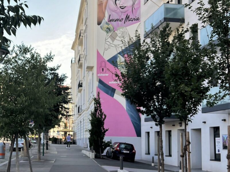 A modern, vibrant mural on a building in Mariahilfer Vienna, with greenery and trees along the sidewalk, offering a mix of contemporary and classic architecture.