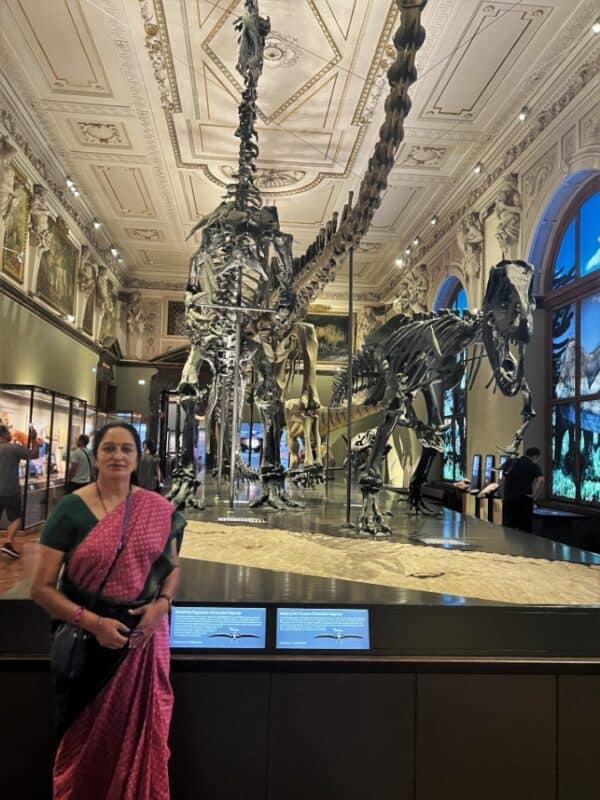 Dinosaur skeletons displayed in a grand gallery with woman in pink saree standing in front at Natural History Museum Vienna