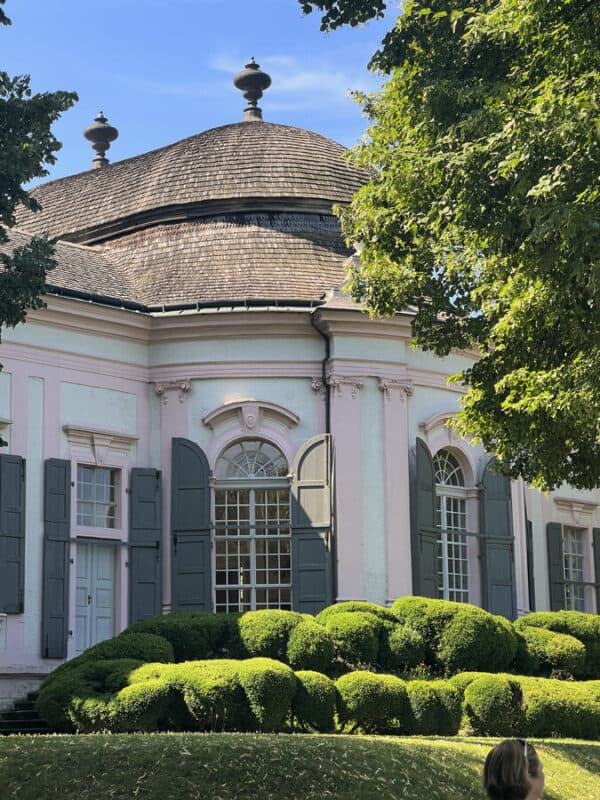 Side view of a pink baroque pavilion with grey wooden shutters and a domed roof, surrounded by neatly trimmed green bushes and leafy trees under blue sky.