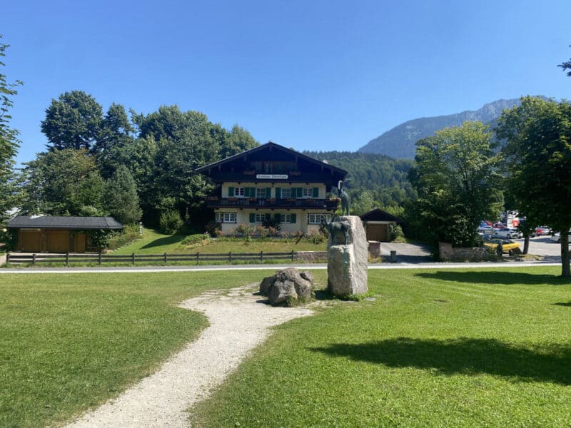 Traditional Bavarian guesthouse in a green park with a statue and surrounding trees near Schönau am Königssee.
