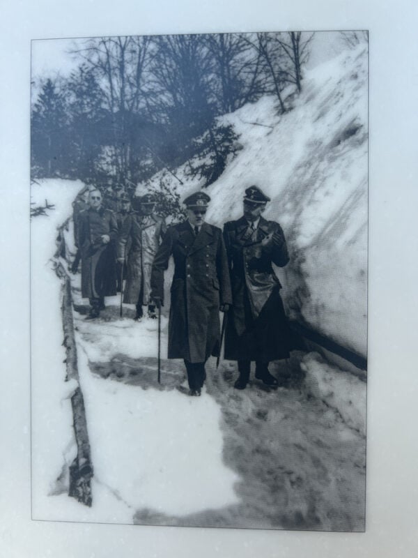 Black and white historical photo showing Adolf Hitler and Nazi officers walking on a snowy path near Obersalzberg
