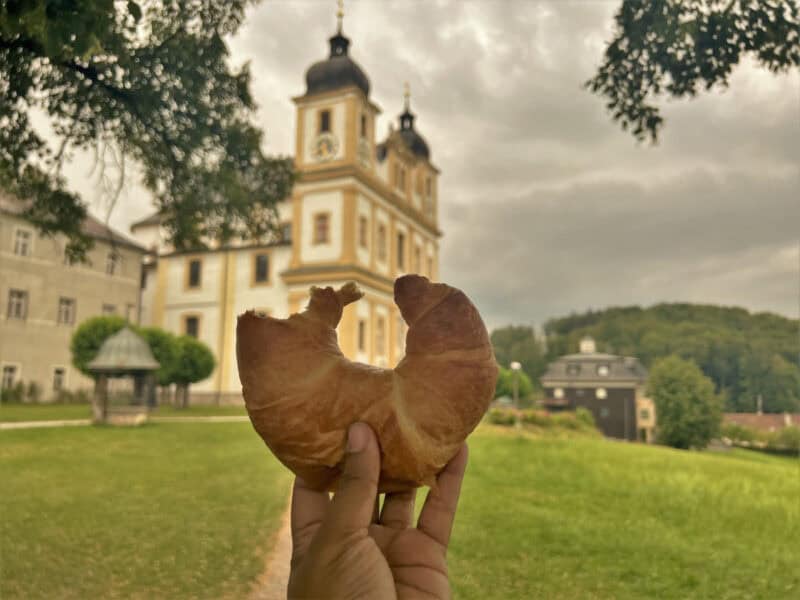Croissant held up in front of Maria Plain church with grassy hill and cloudy sky in background