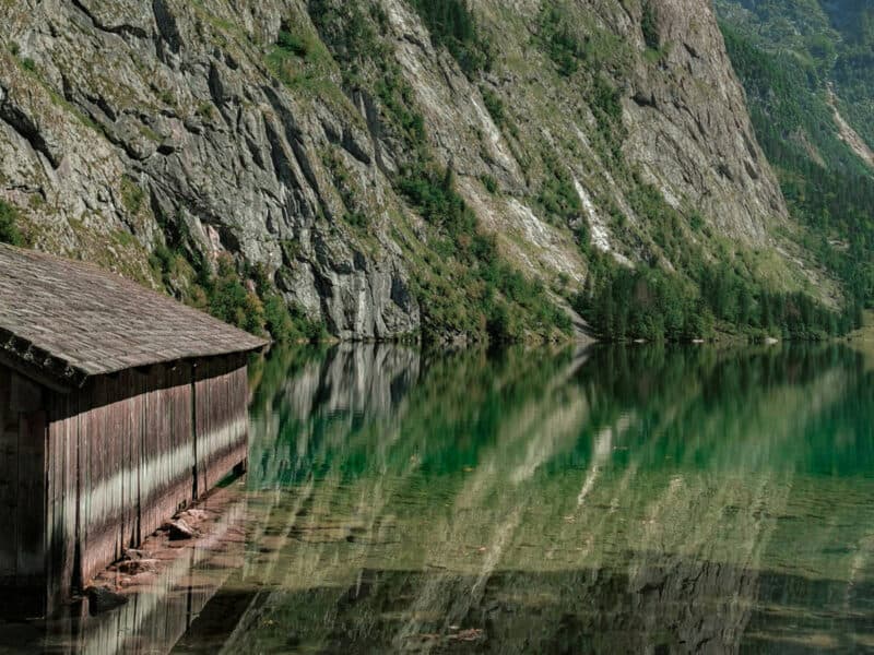 Old wooden boathouse reflected in the still, clear waters of Königssee, with rocky cliffs rising beside the lake